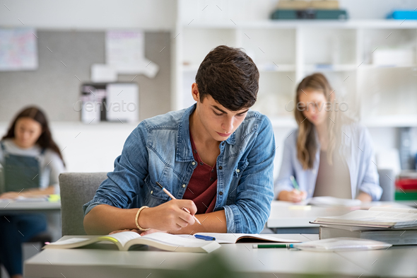 Focused young man taking notes from books for his study. College student sitting at desk with books for finding information in high school library. Guy studying in classroom and completing project with highschool classmates in background.